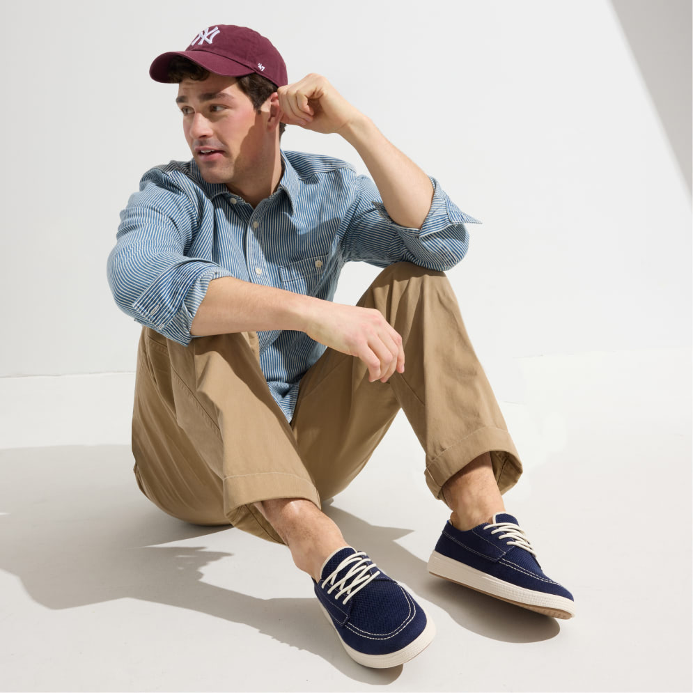 A man in a blue shirt and khaki pants sits on the floor, wearing stylish Ravello boat shoes